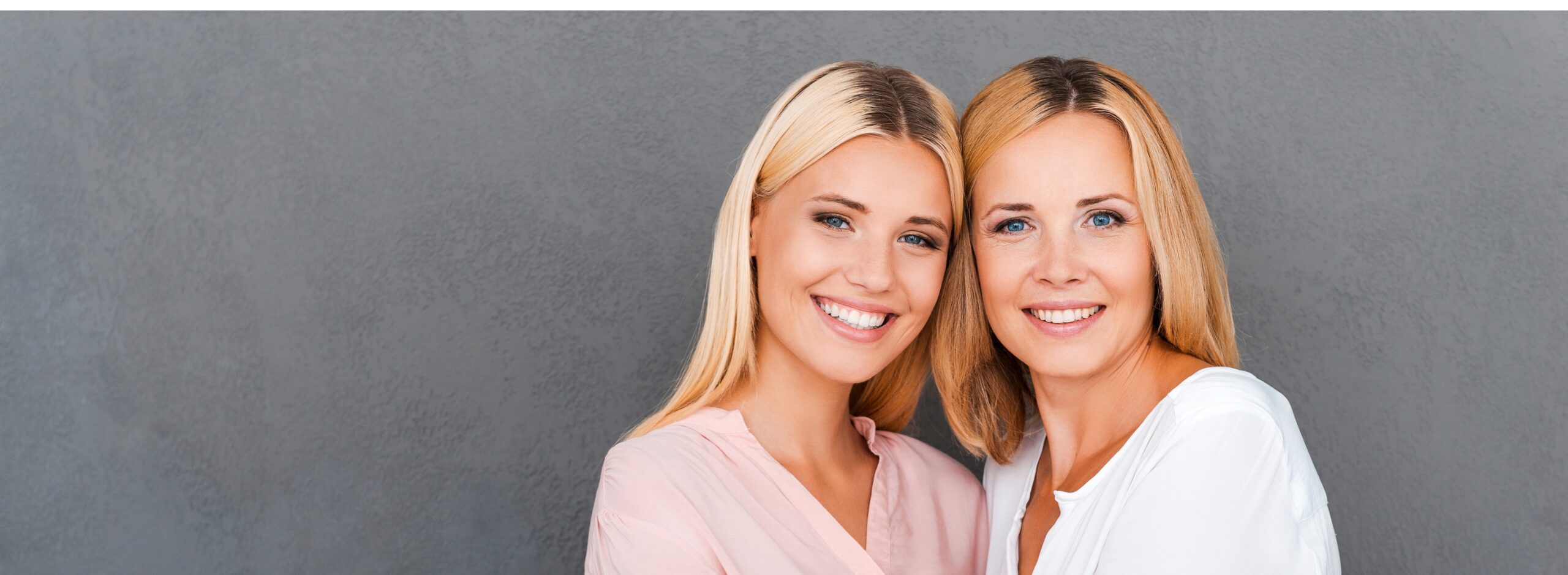 two women with blonde hair smiling and standing against a grey background